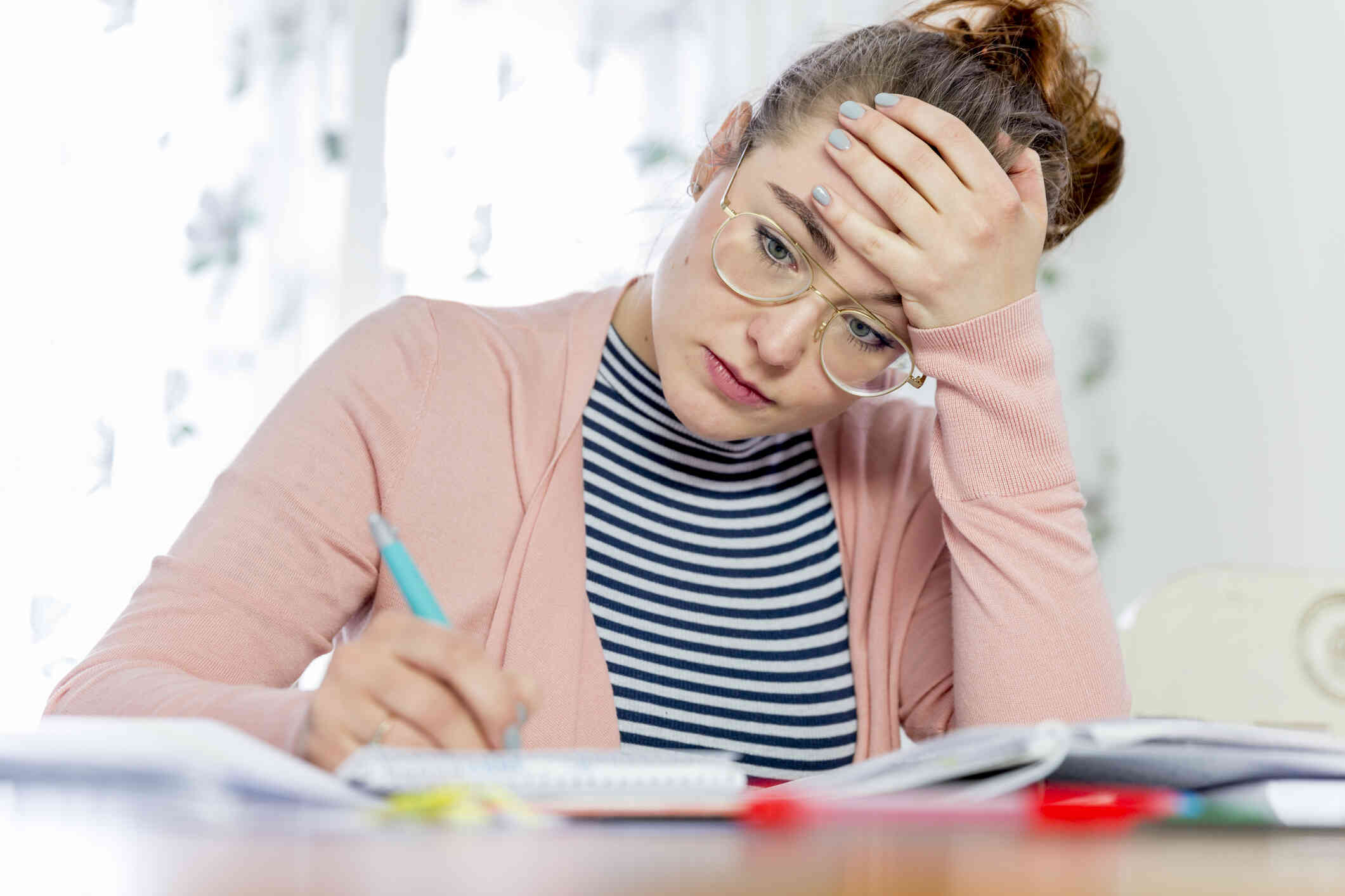 A woman wearing a pink cardigan sits at her desk at work with a worried expression while writting on some papers on the desk infront of her.